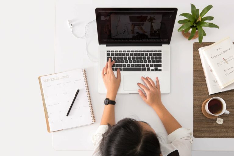 Una donna alla scrivania con quaderno e caffe usa un laptop per cercare un’agenzia per la creazione siti web.