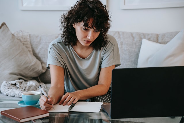 eine Frau, die zuhause auf dem Sofa sitzt, neben sich eine Tasse Kaffee, macht sich Notizen, während sie auf ihrem Laptop eine online-lektion nimmt, um zu lernen, wie sie ihren neuen E-Commerce nutzt