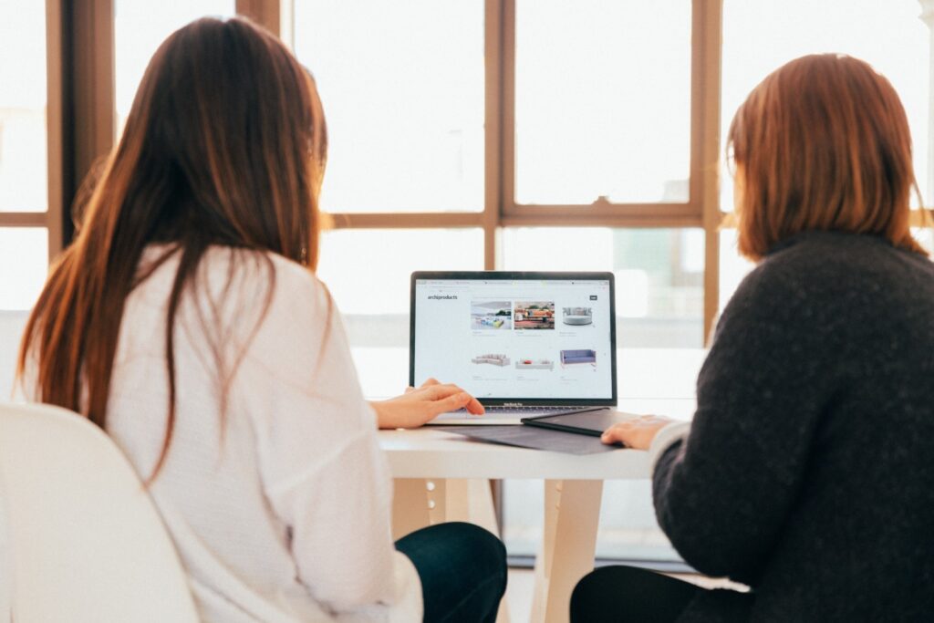 Due donne sono sedute a un tavolo in una sessione di web marketing consulenza e osservano lo schermo di un computer portatile che mostra la pagina di un e-commerce. La consulente web marketing spiega alla sua cliente come può aprire e progettare un e-commerce di successo.